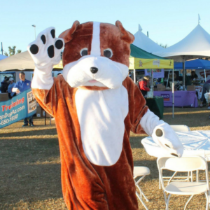 A person in a large dog costume waves with a paw at an outdoor event.