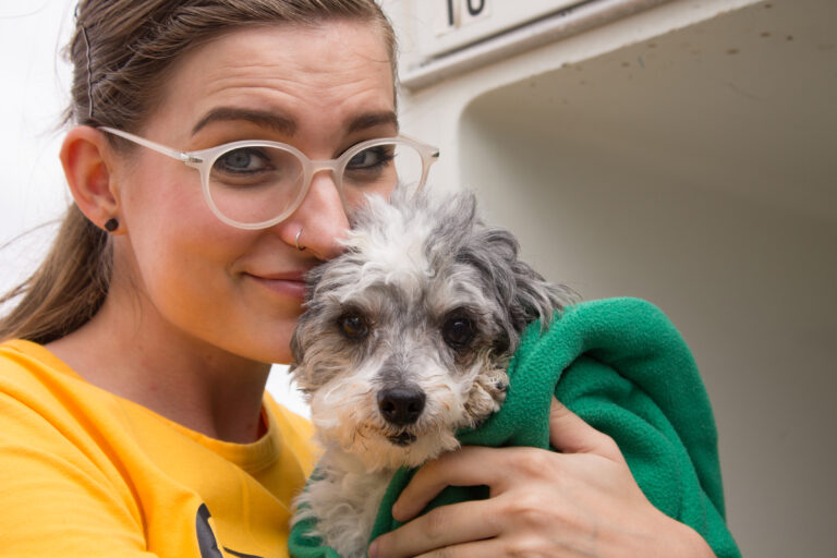A smiling woman wearing glasses cradles a small, wet, fluffy dog wrapped in a green towel.