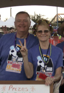 A man and woman pose at an outdoor event wearing matching blue volunteer t-shirts, one giving a peace sign