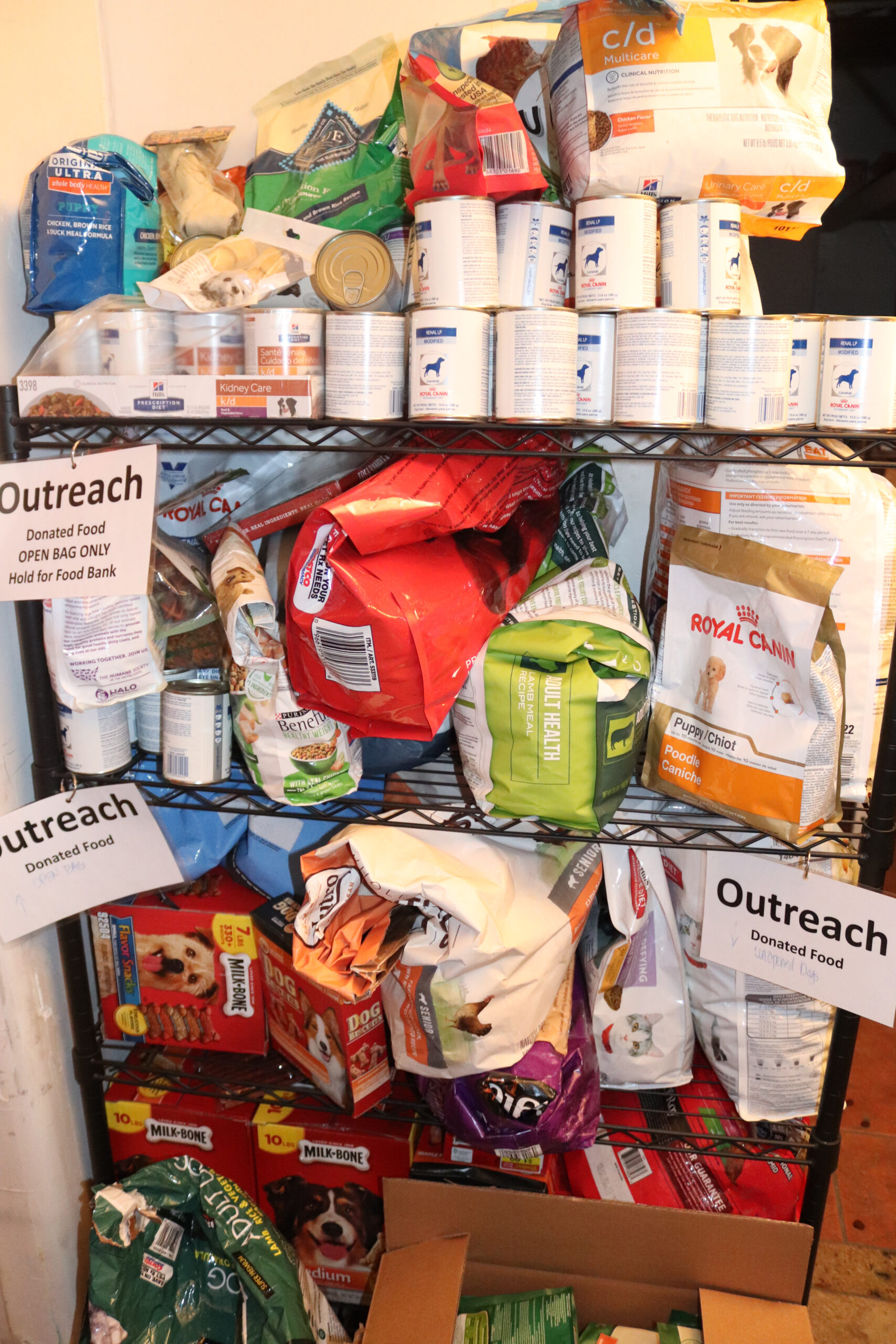 Shelves stacked with bags and cans of donated pet food under signs reading 'Outreach Donated Food'