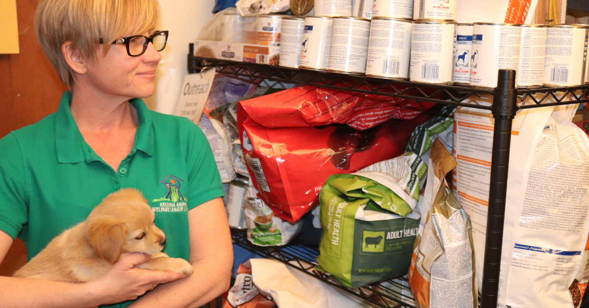 A volunteer in a green polo shirt holds a golden puppy beside shelves of donated pet food