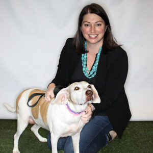A smiling woman kneels and pets a tan and white dog with a purple collar.