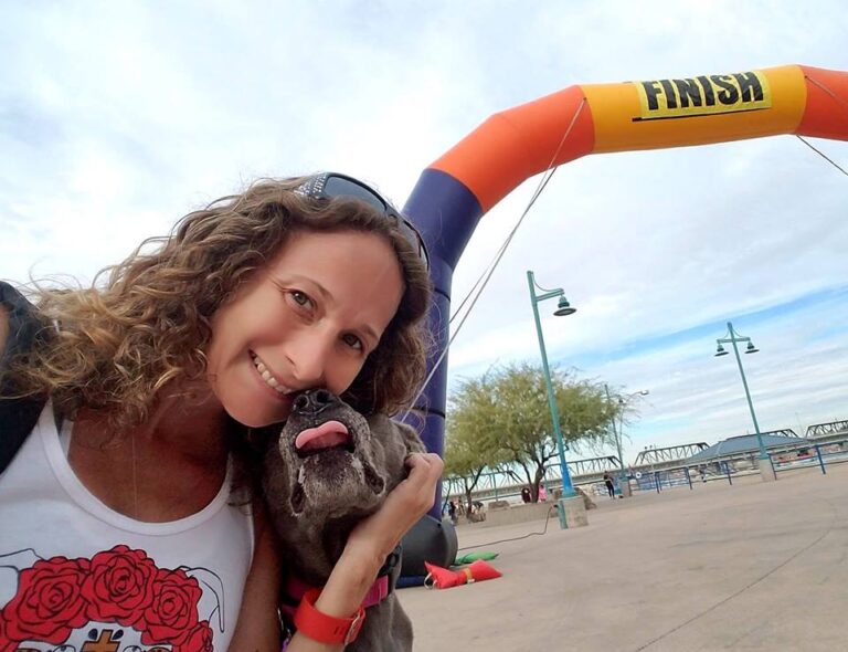 A smiling woman and her dog pose near a race finish line on a sunny day.