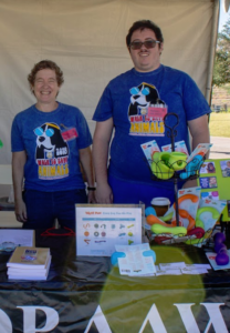 Two volunteers stand behind a table with dog toys and informational flyers at a fundraising event.