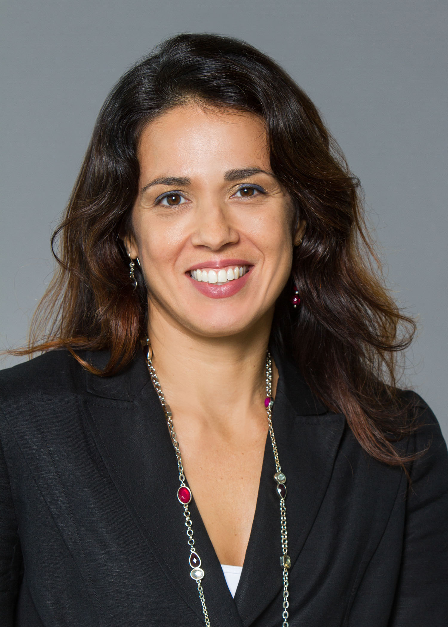 A woman with long brown hair smiles in a professional headshot, wearing a black blazer and a beaded necklace.
