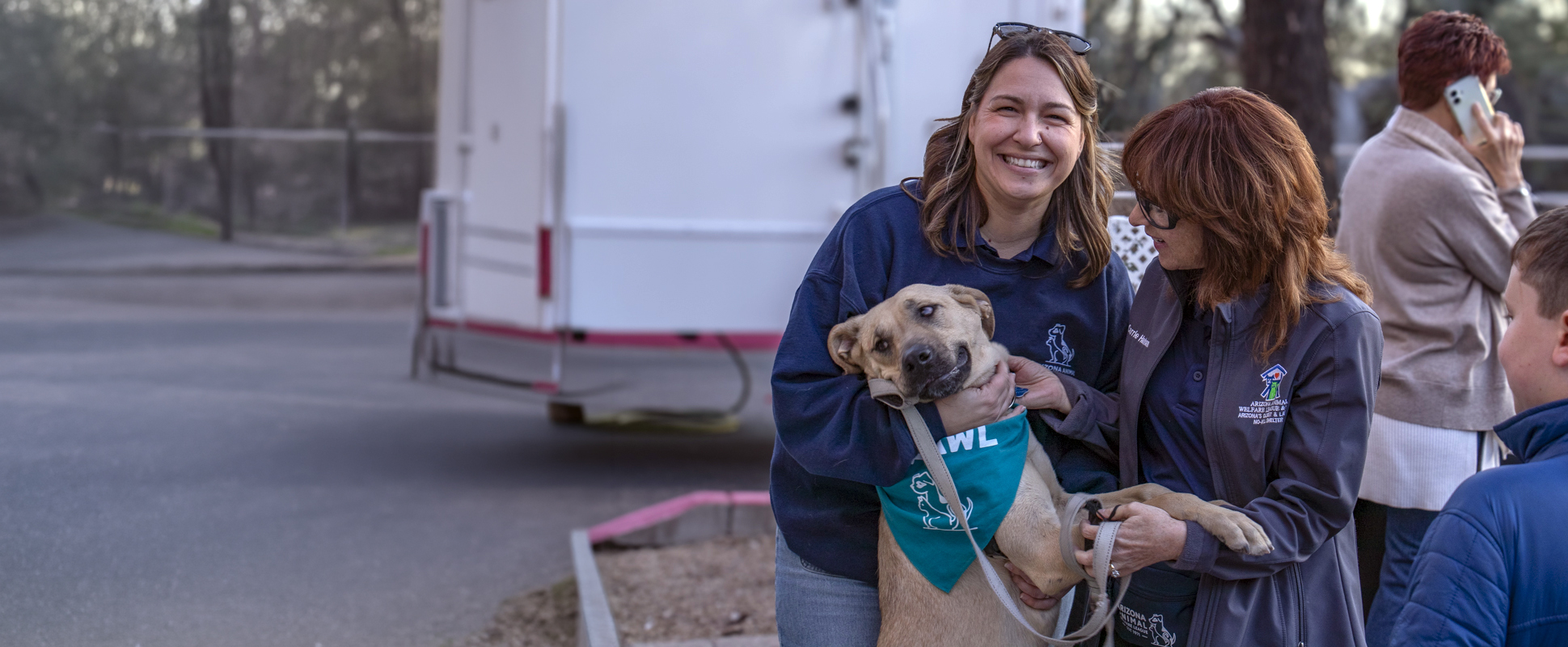 Two staff members comfort a tan dog wearing a teal bandana in front of a mobile clinic trailer