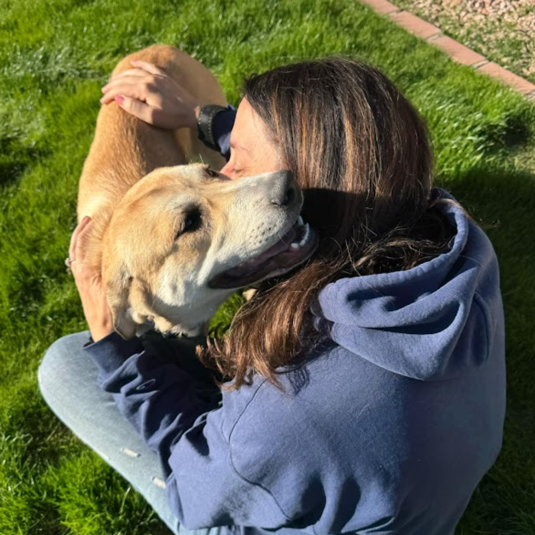 A woman lies on grass hugging and nuzzling a tan dog outdoors