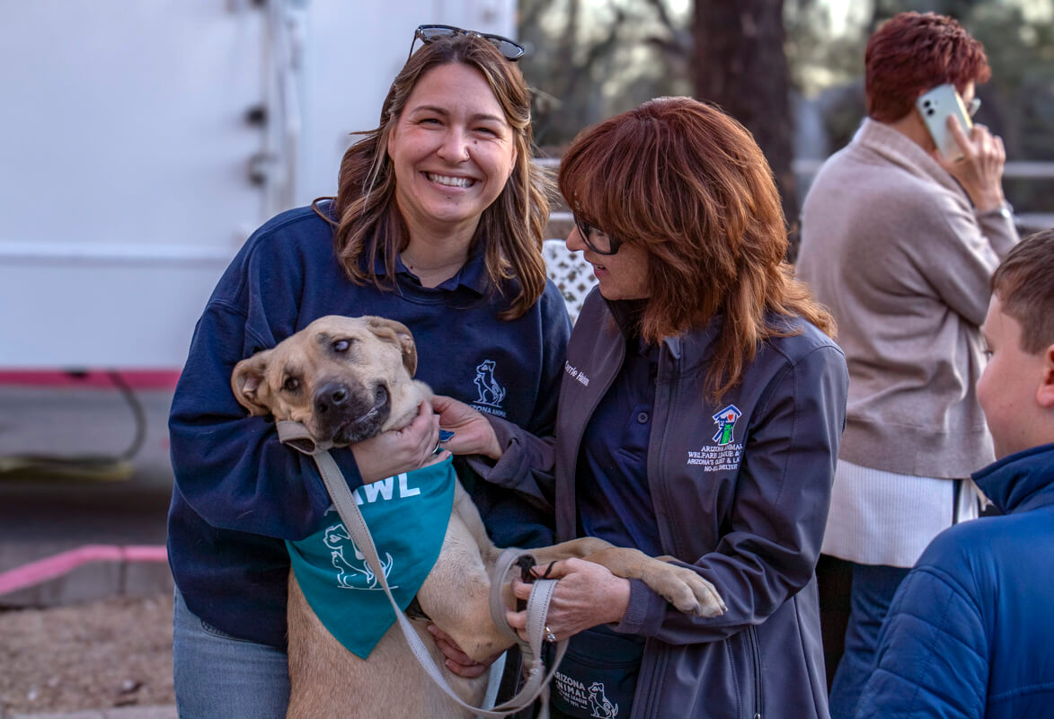Two smiling women hold a senior tan dog wearing a teal AAWL bandana at an outdoor event.
