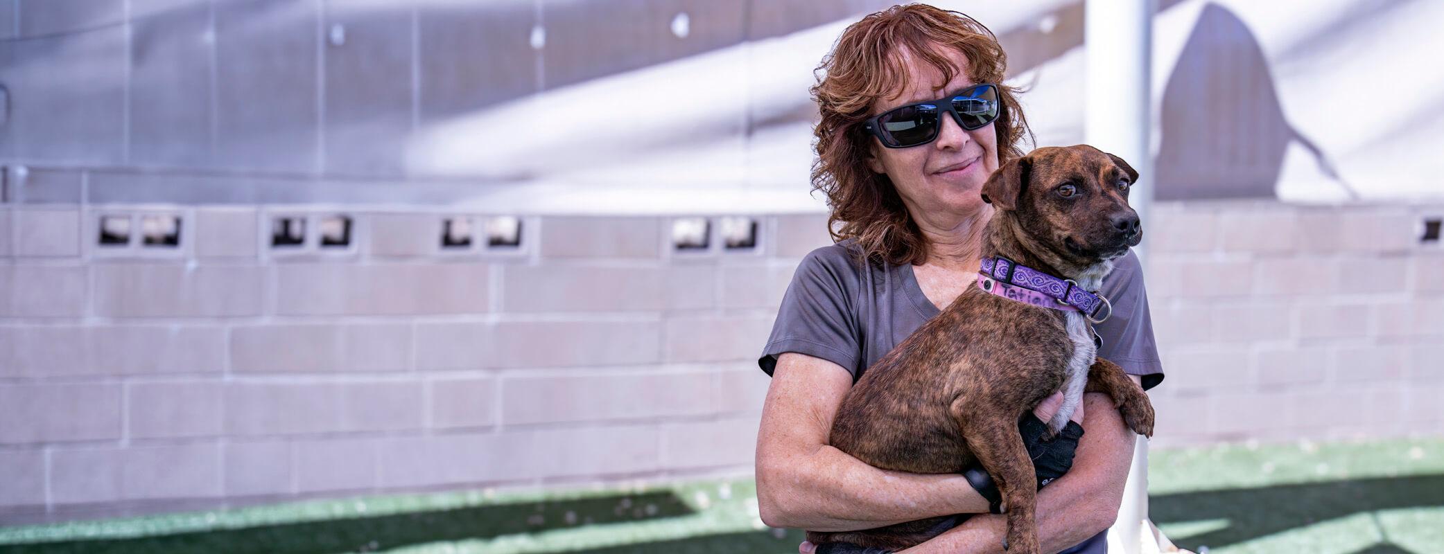 A woman in sunglasses holds a brindle dog wearing a purple collar outdoors.