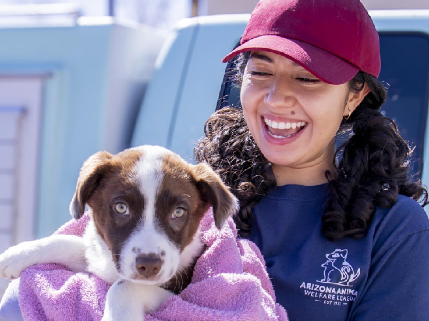 A smiling AAWL staff member in a maroon cap holds a brown-and-white puppy wrapped in a lavender towel beside a mobile clinic van