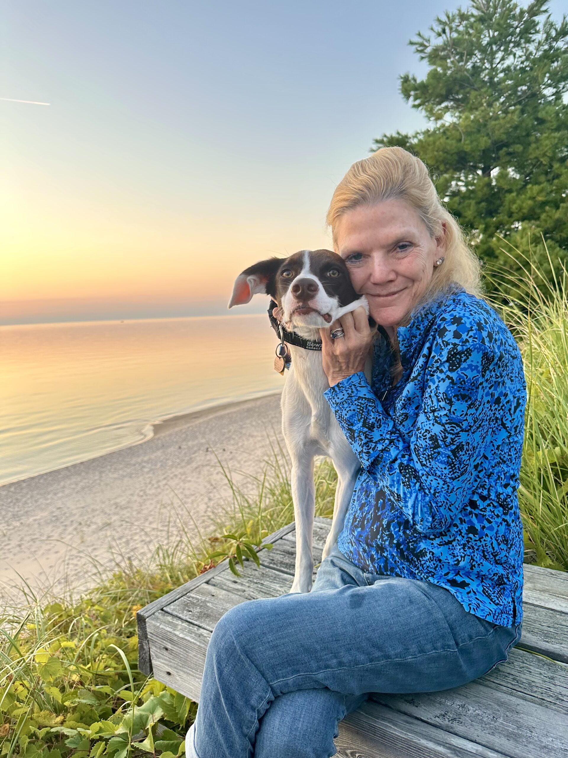 A woman sits on a boardwalk at sunset with her arm around a black and white dog, the beach and water behind them
