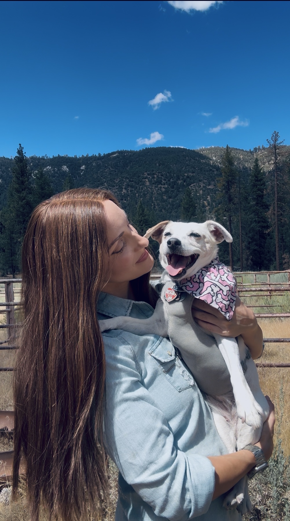 A woman holds a white dog wearing a pink bandana outdoors, with pine trees and mountains behind them