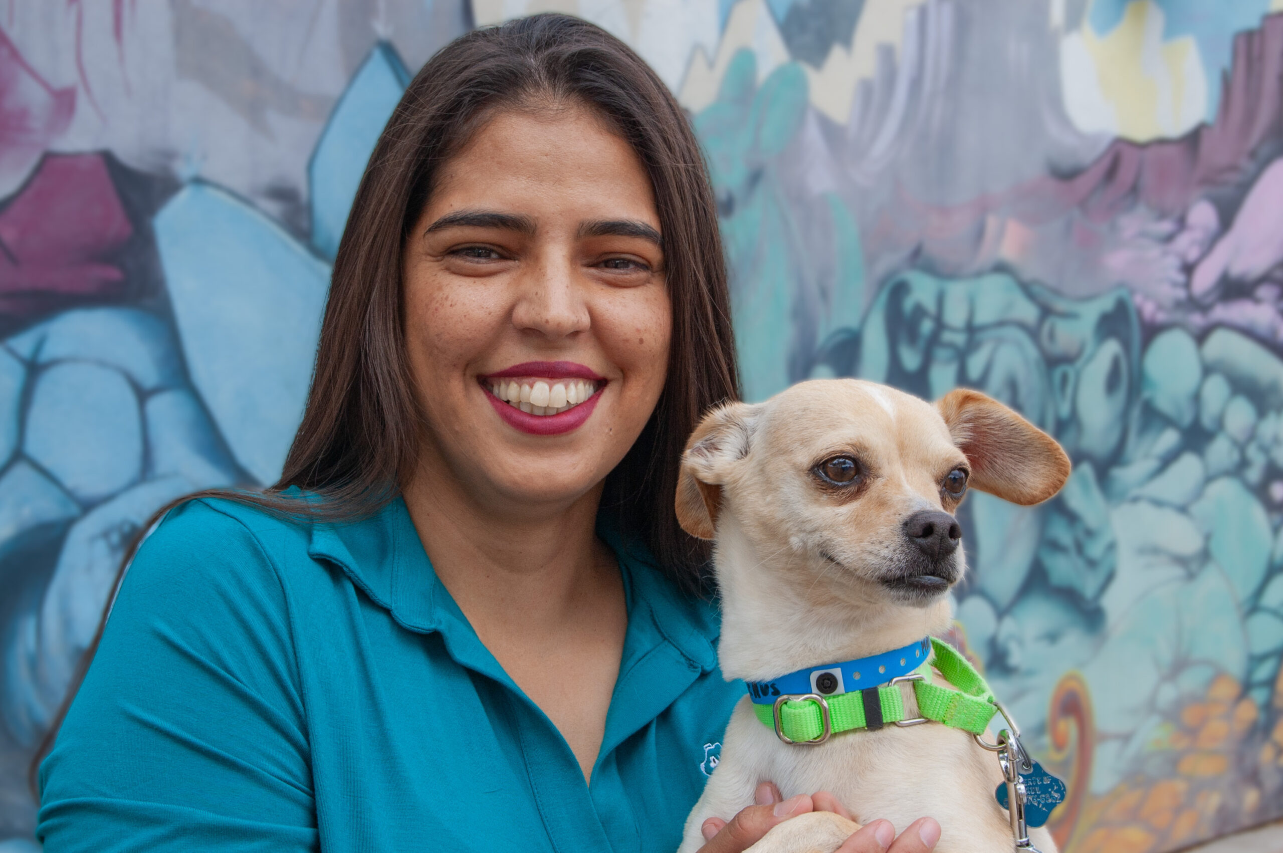 Mujer con polo azul de AAWL sonríe mientras sostiene a un perro chihuahua color crema frente a un mural colorido.