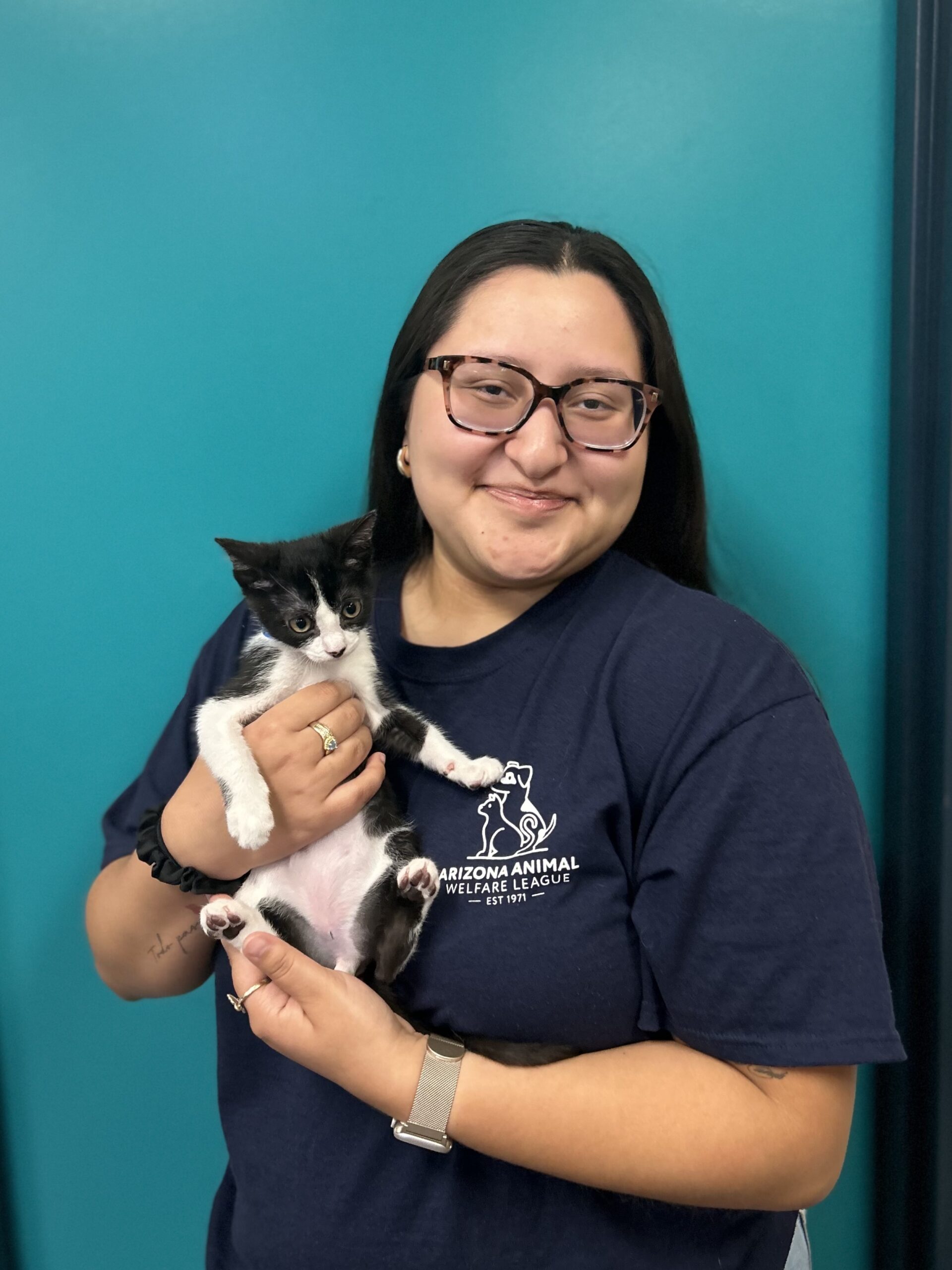 Mujer con gafas de montura de carey y camiseta azul marino de AAWL sostiene a un gatito blanco y negro frente a una pared turquesa.