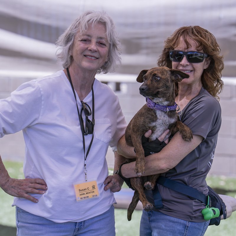 Two volunteers stand together outdoors, one holding a brindle dog in her arms