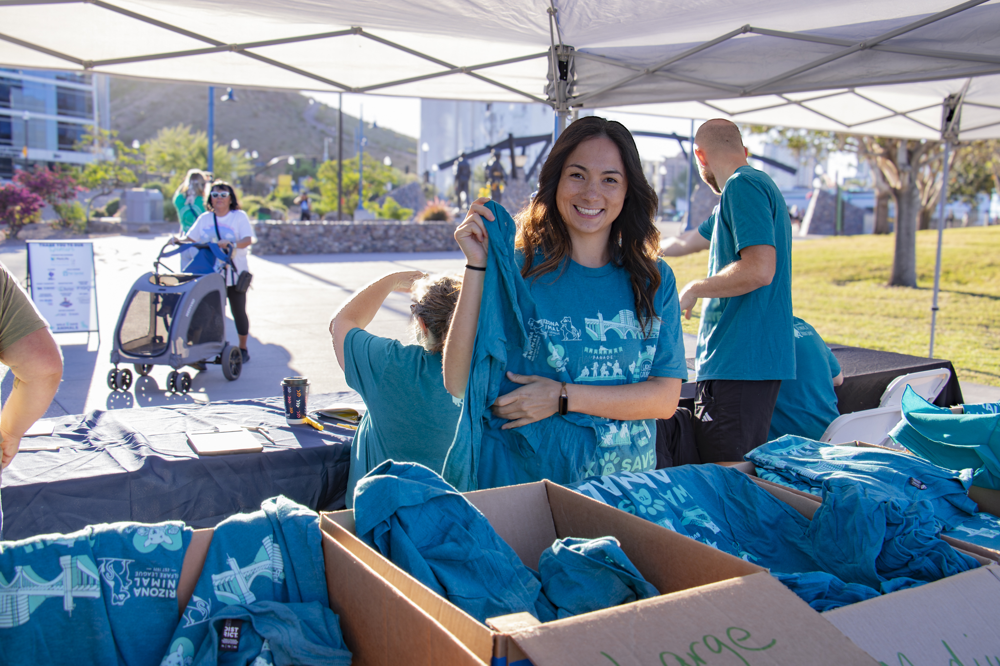 A smiling volunteer holds up a teal AAWL event T-shirt at an outdoor merchandise tent, with boxes of folded shirts on the table beside her
